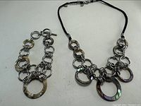 Necklace and bracelet set laid flat showing interlocking rings design with metal and abalone-like shell segments linked by black cord and metal chains