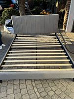 Queen size bed with light gray padded upholstered headboard and footboard, wooden slats for mattress support, and black metal frame.