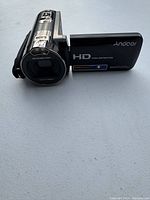 Front/angled view of Andoer HD handheld video camera showing lens, flip-out screen with branding, and side hand strap.