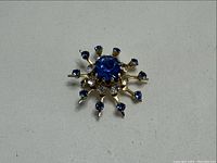 Close-up photo of vintage Coro brooch with central large blue rhinestone and surrounding smaller blue and clear rhinestones in a gold-tone sunburst setting.