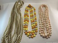 Photo showing three vintage costume necklaces laid side by side: cream and brown beaded necklace, orange/yellow bead necklace, pinkish pearl-like bead necklace.