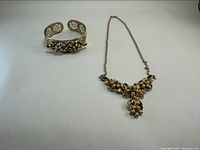 Photo showing the vintage necklace with a floral cluster pendant and matching open cuff bracelet with floral and pearl details laid flat on white background.