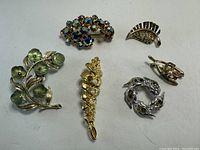 Photo showing six vintage pins and brooches with floral and decorative designs in various metal tones and stones.