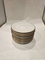 Stack of 11 small white porcelain plates with gold gilded rims and center ring, showing front top profile.