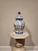 Front view of an antique blue and white ceramic ginger jar vase with matching lid topped with a blue finial, placed on a decorative pedestal.