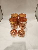 Four amber glass cups with grape and leaf embossed patterns grouped together with two small amber glass floral frog vases in front, all against a white background.