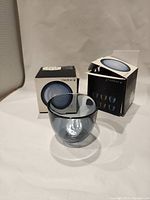 Two Orrefors Pastillo steel blue glass bowls displayed with their original boxes behind each bowl, showing open box flaps and the bowls in front.