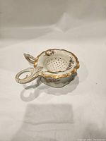 Porcelain tea strainer side view showing scalloped edge, gold trim, floral design, and curled handle.
