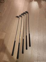 Five Prima golf irons laid out on a wooden surface showing full length of clubs with black grips and metal shafts.