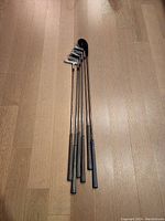 Photo showing 5 golf clubs with metal heads and grips, laid flat for condition and count assessment.