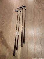 Four Tommy Barton Signature golf irons laid out on a wood floor showing full length of clubs including grips.