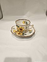Tea cup and saucer with yellow flowers and gold edging on white bone china surface.