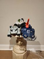 Three vintage action figure vehicles displayed on a pedestal with a beige wall background: a blue Thundertank with orange cannon, a white and green tank, and a white and black vehicle.