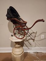 Side view of the wicker wood doll pram showing the folded-down black canvas hood, wooden handle, and metal bands on wheels.