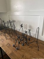 Photo showing 10 black metal display stands of varying styles and heights arranged on a wooden table against a white wall.