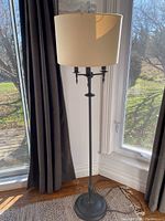 Full view of the black spray painted floor lamp standing next to windows with grey curtains, showing the entire height and beige lampshade.