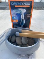 Photo showing Conair Extreme Steam handheld fabric steamer box with image and product claims, next to ceramic bowl with smooth massage stones and wooden tongs.
