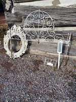 Overall lot including metal headboard, plastic white frames, birdhouse-shaped thermometer on stake, and metal wire planter on gravel in front of wooden background.