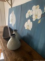 Photo showing two orchid paintings leaned against wall, two ceramic vases in front, and narrow wood server surface.