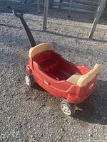 Full view of red plastic two-seat wagon outdoors on gravel surface showing handle, seats and wheels