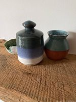 Three handmade pottery pieces placed on a wooden surface against a white wall, including a lidded jar, a vase, and a small bowl partially visible.