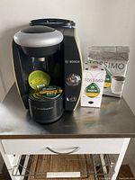 Full front view of Bosch Tassimo machine with coffee discs and a white bag of Tassimo coffee discs on stainless steel surface.