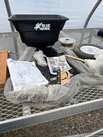 Various parts of the Blue Hawk tow-behind spreader laid out on a metal table including black hopper, frame parts, wheels, hardware, and manual.