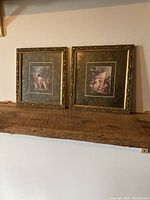 Two vintage framed prints with gold ornate frames and dark mats on wooden shelf against wall.