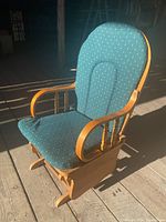 Side angled view of solid wood glider rocking chair with teal polka dot upholstered cushions on seat and backrest, visible armrests and rounded wooden base.