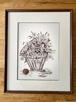 Full view of the framed needlepoint artwork showing the cat in a flower basket with flowers and foliage, neutral background, wooden frame with minor wear.