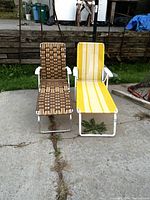 Two vintage folding loungers placed side by side on concrete, one with brown woven webbing and the other with yellow striped fabric, showing slight rust.