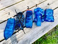 Four cobalt blue glass Avon cologne bottles shaped as one boat and three cars, arranged linearly on wood surface.