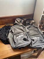 Photo showing three identical gray Metis backpacks with white floral logos and text on front pockets, and one older brown and black duffel bag behind them on a wooden surface.