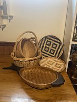 Photo showing five woven baskets and trays arranged on surface, with two handled baskets, two patterned trays, and one shallow tray with a chip.