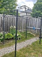 Full view of the black metal BBQ pergola frame set on grass beside a wooden fence, showing the tubular metal legs and peak roof structure. Also includes a black metal shelf in background not included with the lot.