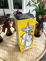 Four copper-tone metal Mexican musician sculptures each with a traditional hat and string instrument, standing on a woven tray, with original yellow and black box visible.