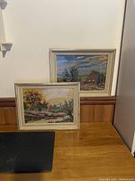 Two hand embroidered landscape pieces in distressed off-white frames resting against a wall on a wood surface.