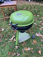 Green kettle-style charcoal BBQ grill outdoors on grass, showing slight rust and vent on lid