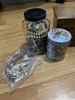 Photo showing a large glass jar filled with assorted jewelry and beads, a decorative tin container with floral design, and a sealed plastic bag with white and brown beads and jewelry pieces on wooden floor.