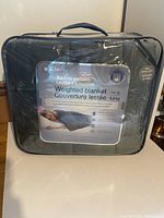 Front view of weighted blanket packaged in a sealed transparent carrying case showing brand, product details, and imagery.
