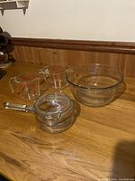 Lot of four glass kitchen items: two Pyrex measuring cups with red markings, one large Anchor Hocking mixing bowl, one Pyrex lidded saucepan. All items on wooden countertop against wood paneling.