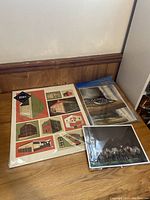 Top view of three art print sets: hand screen printed "7 Sheep" pastel print by Jane MacMillan, set of 8 laminated watercolor prints by Andrew P. Woodhouse, and a hand screen printed poster of buildings.