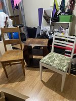 Photo showing two chairs beside a vintage magazine side table with visible wear and needing refinishing. The natural wood chair has a simple ladder back design, and the white chair has a green leaf patterned cushion.