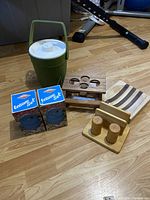 Overview of the vintage camping lot showing the green plastic ice bucket with white lid and handle, wooden salt and pepper shakers on tray, two unopened boxed Leisure Set camping items, a wooden holder with four round holes, a wooden condiment holder, and a striped wooden tray placed on a wood floor.