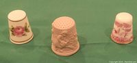Three thimbles displayed side by side on green fabric showing individual decoration: Spode pink rose, Royal Worcester unglazed pottery with figures, China thimble with pink Mill Pond Scene.