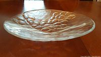 Side view of large clear glass platter with raised grape cluster design showing heavy, thick glass and slightly uneven edges.
