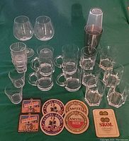 Photo showing all items in lot arranged on green cloth: 2 Pinot Noir wine glasses, 4 beer mugs, 6 rock whiskey glasses, 6 stackable whiskey glasses, vintage cocktail shaker, 8 bar coasters.