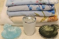 Stack of towels showing white bath sheet on bottom and floral patterned bath towels above, with white and chrome toothbrush holder in foreground, and two plastic clamshell soap dishes in blue and olive placed in front.