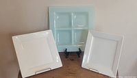 Front-facing image of the light blue 4-compartment glass tray centered between two white square ceramic plates on display stands.