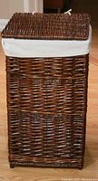 Front and side view of a rectangular dark brown rattan laundry hamper with hinged lid and removable beige linen liner visible at the top edge.
